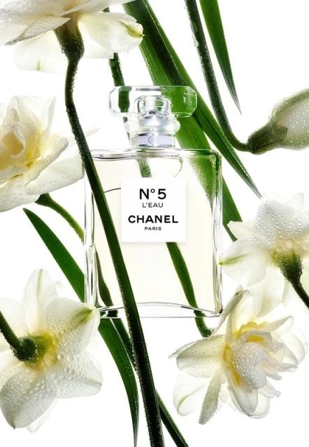 Close-up of a Chanel No. 5 L'Eau perfume bottle surrounded by white daffodils with water droplets and green stems against a white background.