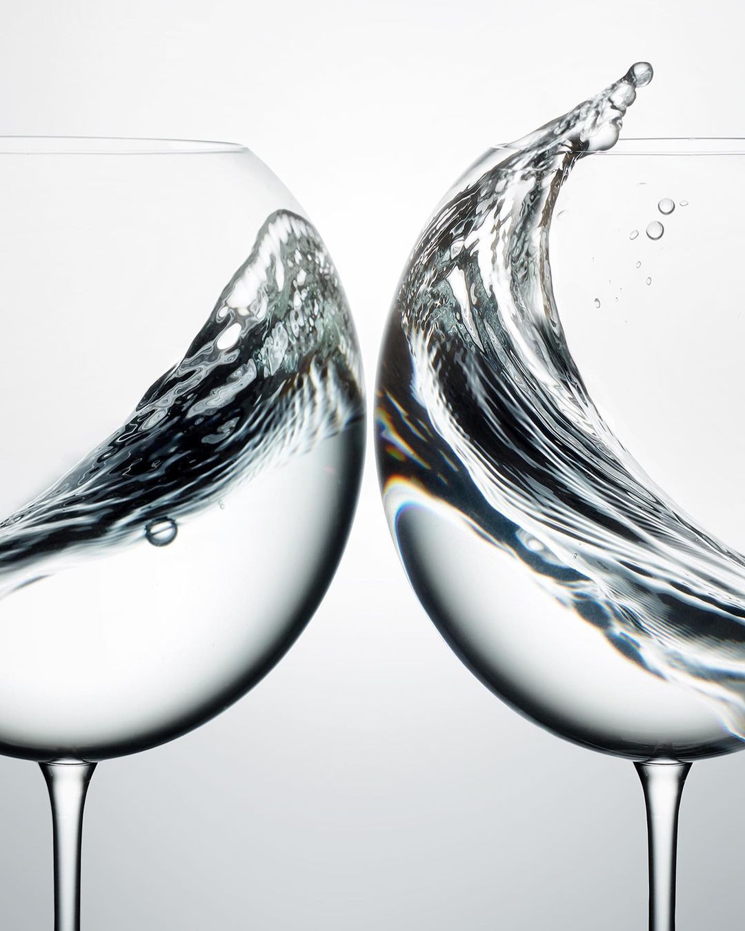 Two wine glasses collide, water splashing in mid-air against a bright background, capturing a dynamic moment in a studio environment.