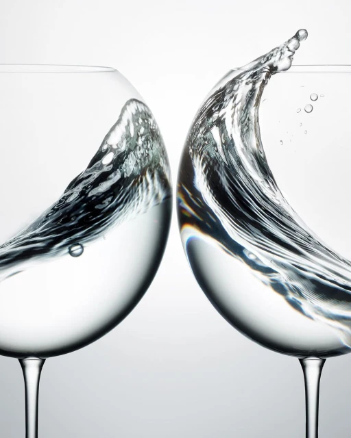 Two wine glasses collide, water splashing in mid-air against a bright background, capturing a dynamic moment in a studio environment.