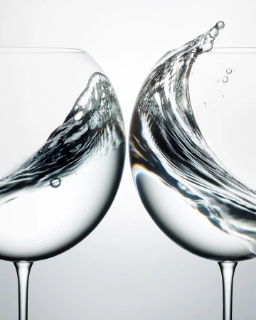 Two wine glasses collide, water splashing in mid-air against a bright background, capturing a dynamic moment in a studio environment.