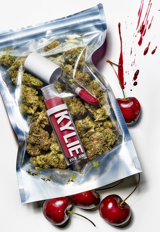 Close-up of a lip gloss and a transparent bag filled with cannabis buds next to several cherries, artfully arranged on a white background.