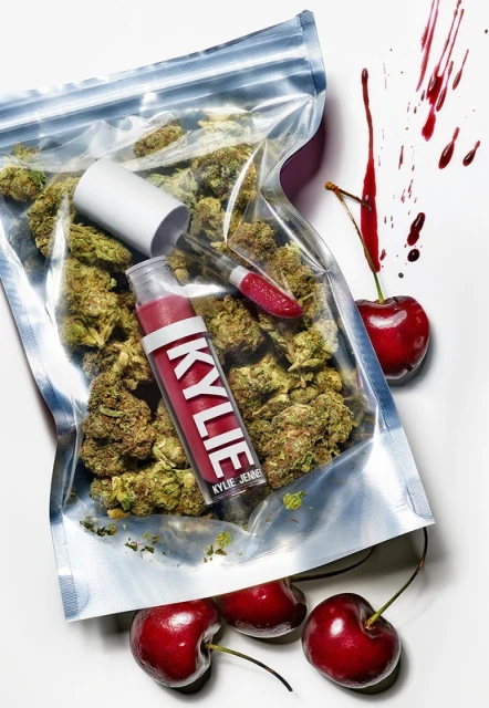 Close-up of a lip gloss and a transparent bag filled with cannabis buds next to several cherries, artfully arranged on a white background.