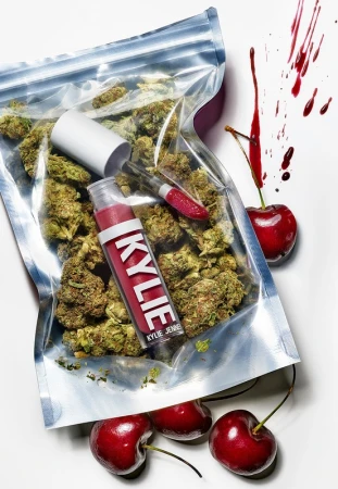 Close-up of a lip gloss and a transparent bag filled with cannabis buds next to several cherries, artfully arranged on a white background.