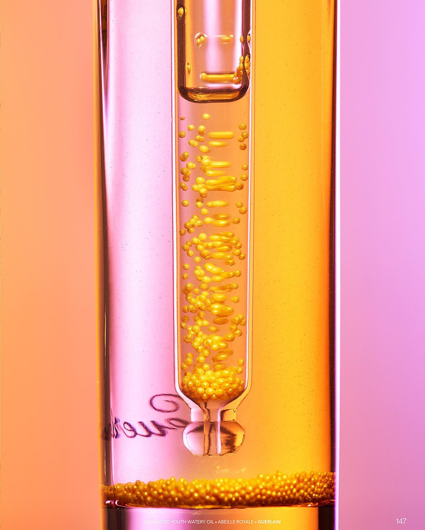 Close-up studio shot of clear glass test tube with pipette containing golden liquid droplets, set against pink and yellow gradient background.
