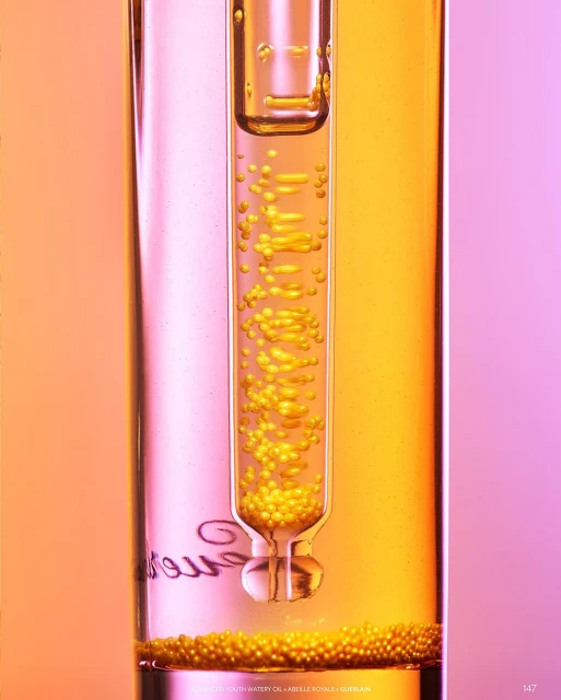 Close-up studio shot of clear glass test tube with pipette containing golden liquid droplets, set against pink and yellow gradient background.