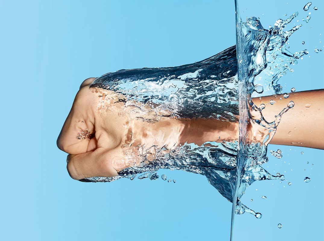 Close-up shot of an adult hand breaking through water, with splashes and bubbles against a light blue background. Aquatic action.