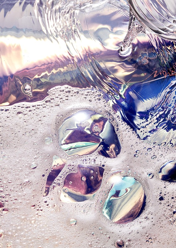 Abstract close-up of iridescent liquid with soap bubbles and reflections, showcasing pastel hues of blue, purple, and cream.