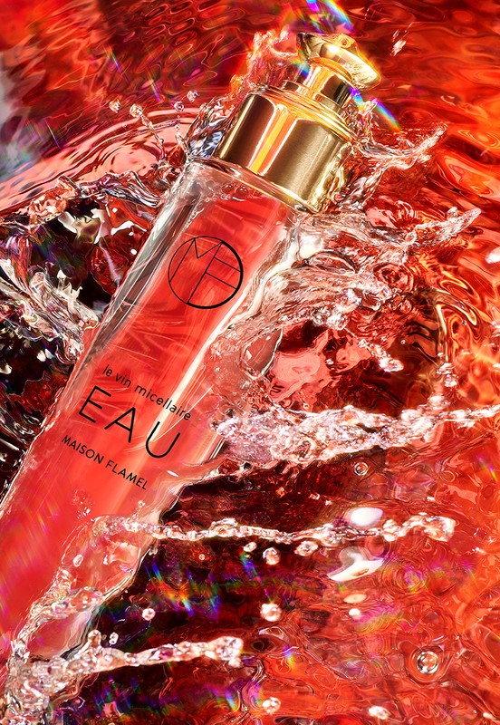 Elevated view of a clear bottle of red liquid with a gold cap amidst splashing water against a blurred red background.