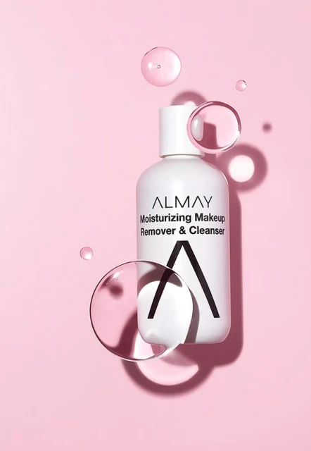 Almay moisturizing makeup remover and cleanser bottle sits on a pink background with several clear, spherical water droplets.