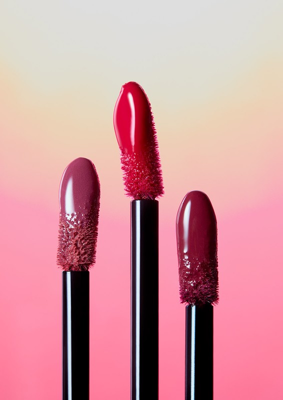 Close-up of three lipstick wands with glossy product on a pink and cream gradient background. Beauty and makeup product photography.