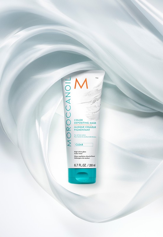 Moroccanoil Color Depositing Mask in white tube with turquoise cap surrounded by light blue swirling silk on white backdrop.
