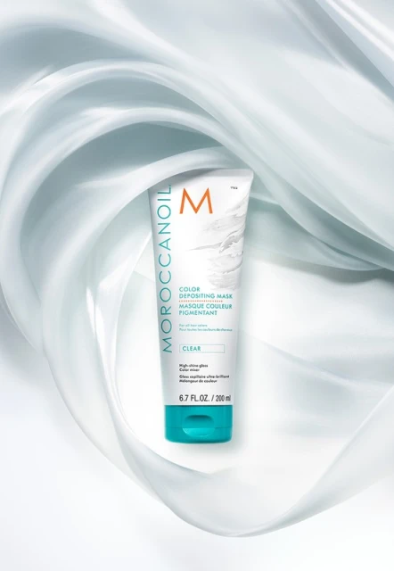 Moroccanoil Color Depositing Mask in white tube with turquoise cap surrounded by light blue swirling silk on white backdrop.