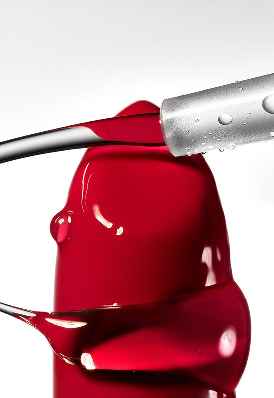 Close-up of red lipstick with a brush applicator, showcasing the texture and color against a clean white background in studio lighting.