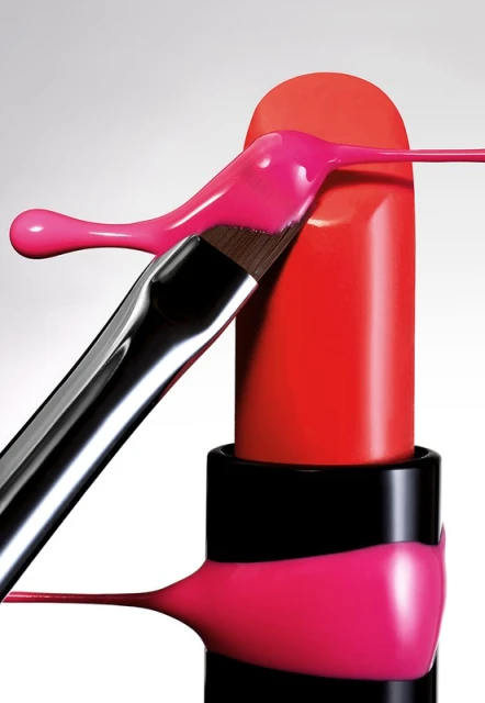 Close-up of vibrant red lipstick with pink paint dripping down its side, applied with a makeup brush, set against a neutral backdrop.
