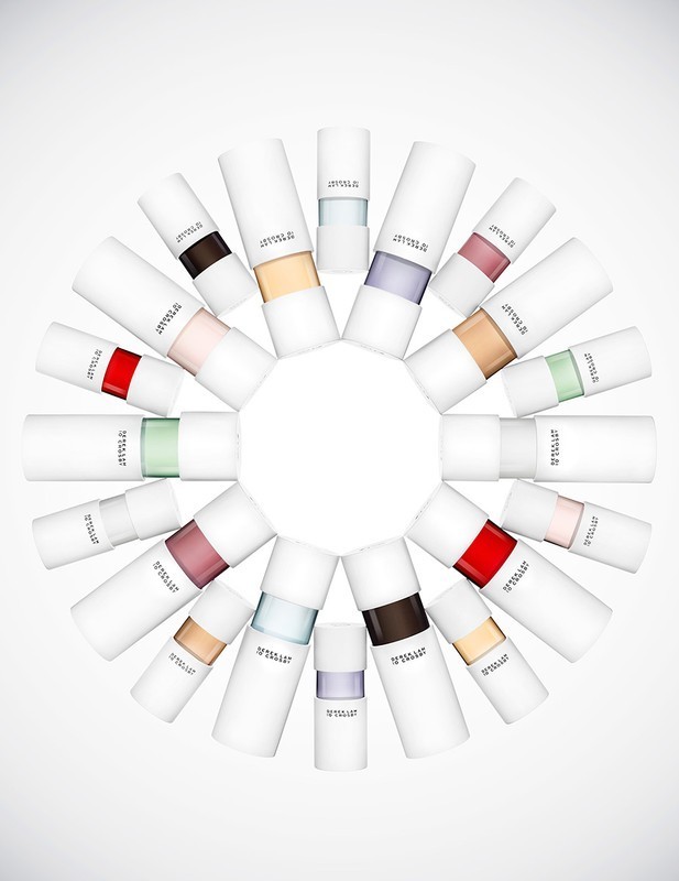 Arrangement of twenty-one white cylindrical cosmetic tubes, each with a different band of color, on a gradient white backdrop.