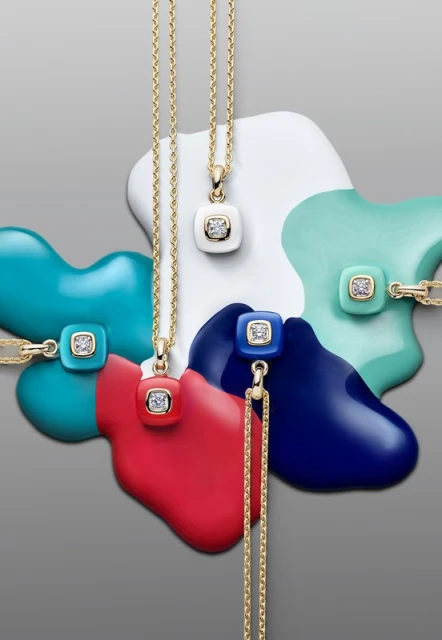 High-angle studio shot of gold chain necklaces with colorful square gemstone pendants against a neutral grey background with glossy paint swirls.