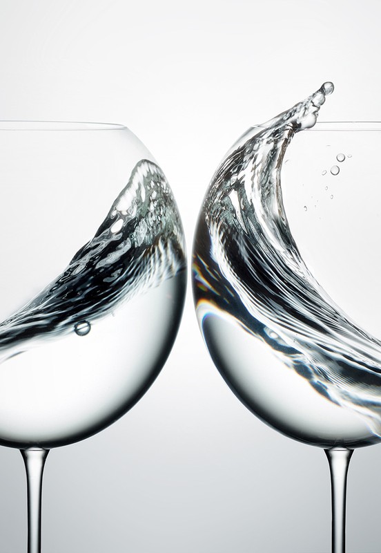 Two wine glasses filled with water capturing dynamic splashes and ripples inside against a clean light background.