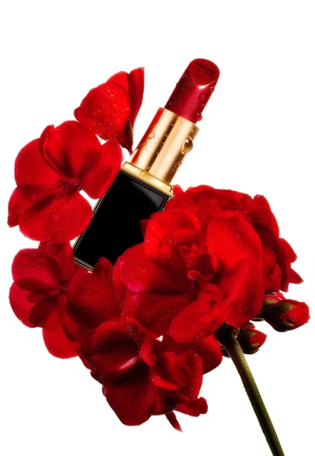 Close-up of red lipstick with black and gold tube surrounded by vibrant red flower petals and water droplets on a white background.