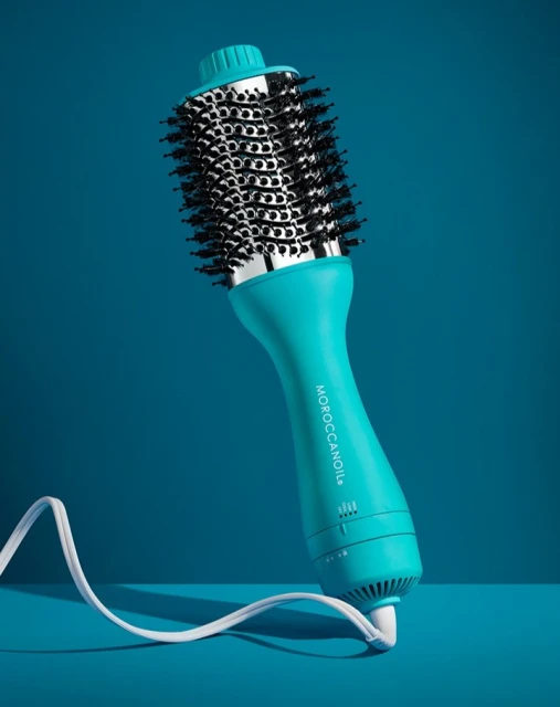 Turquoise hot air brush against a matching teal background. A white power cord is coiled on the surface in front of the hair tool.
