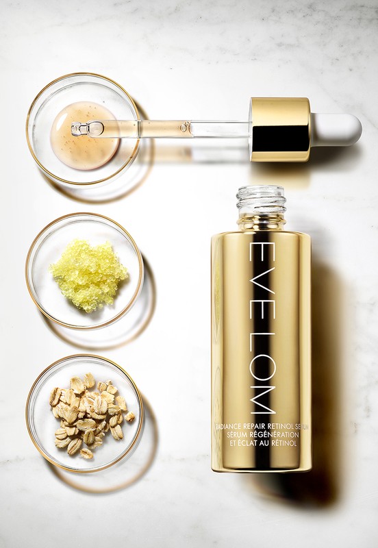 Golden serum bottle with pipette applicator and small bowls of raw ingredients displayed on a light marble surface in studio setting.