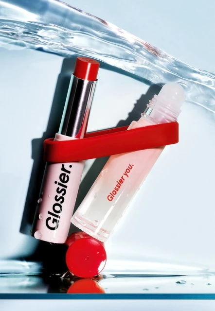Two tubes of Glossier lip gloss and balm are suspended in clear liquid with a light blue background, creating a beauty product still life.