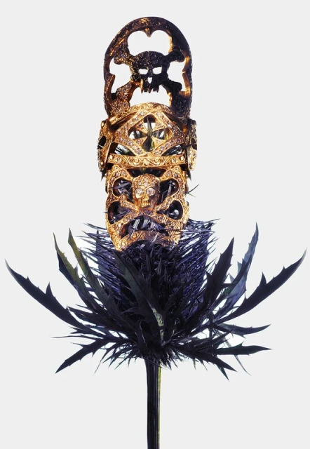 Ornate thistle plant with gilded skulls and crossbones sculpture on top, set against a plain white background. Gothic and intriguing.