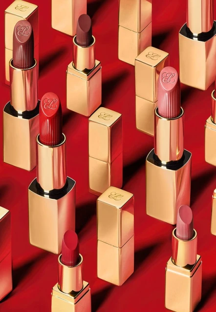 Several gold lipstick tubes arranged in a pattern on a red background with various shades of lipstick on display on some of the tubes.