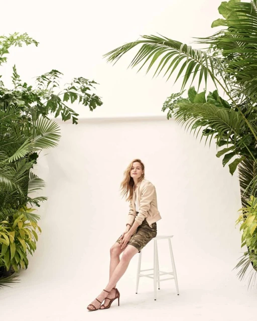Adult woman sits on a white stool in front of a white background surrounded by green tropical plants wearing a beige jacket and camo shorts.