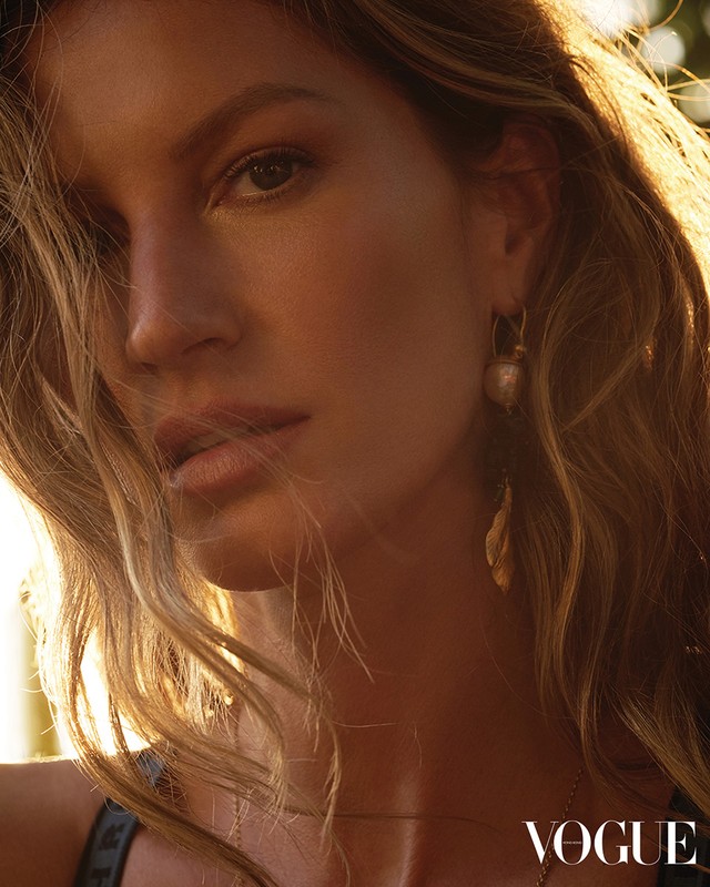 Close-up portrait of a blonde woman with sunlit hair and golden earrings, posing with a serious facial expression. Vogue logo visible.