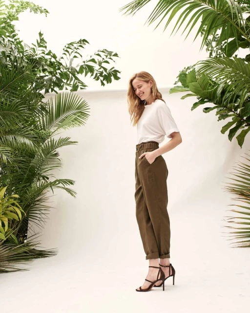 A slender woman in a white t-shirt and olive trousers stands in front of green plants against a bright white backdrop, smiling slightly.