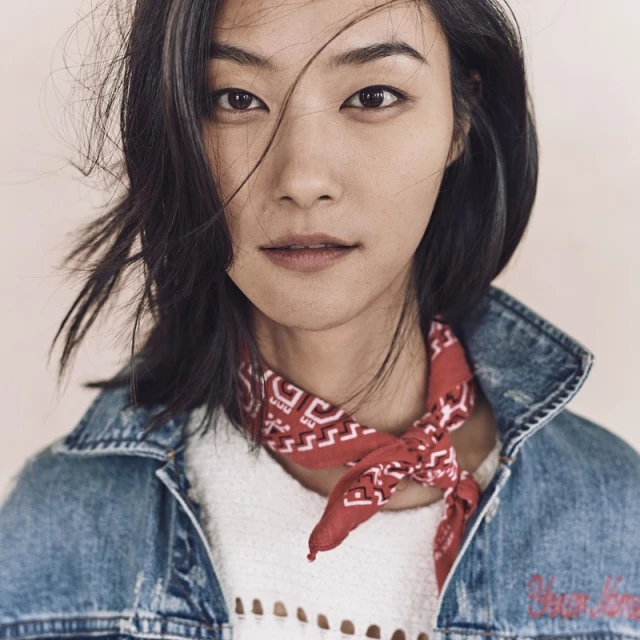 Close-up portrait of a young Asian woman with dark hair, wearing a denim jacket and red patterned bandana, posing against a neutral backdrop.