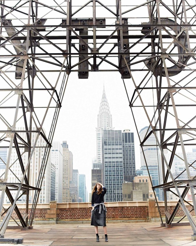 Woman standing on a rooftop with the Chrysler Building in the background framed by a metal structure. Urban cityscape, daytime.