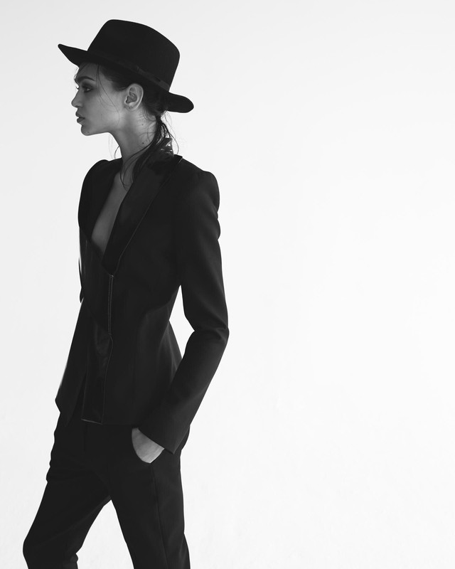 Monochrome portrait of a woman in profile wearing a black fedora hat and tailored dark suit against a plain white background.
