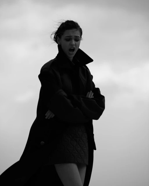 Monochrome portrait of a young woman in an overcoat with crossed arms, against a bright sky backdrop, expressing a strong emotion.