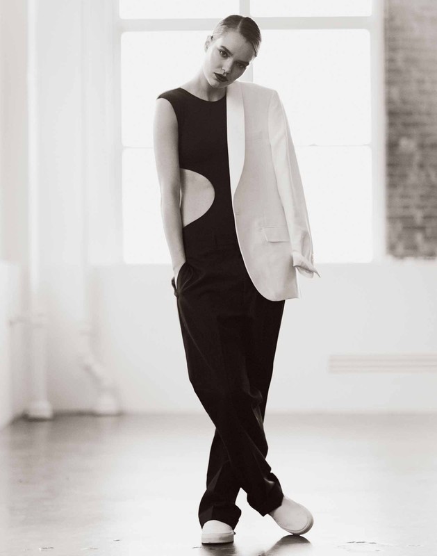 Model in black cut-out top and trousers with a white blazer draped over one shoulder in a bright interior space.
