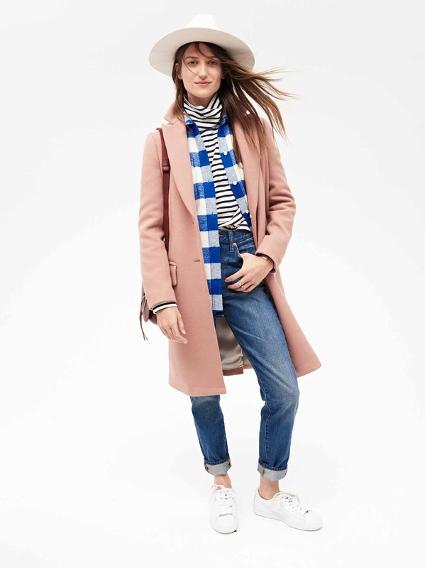 A stylish woman in a salmon pink coat, blue jeans, and a white hat stands confidently against a bright white backdrop, showcasing modern fashion.