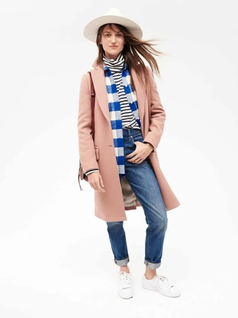 A stylish woman in a salmon pink coat, blue jeans, and a white hat stands confidently against a bright white backdrop, showcasing modern fashion.