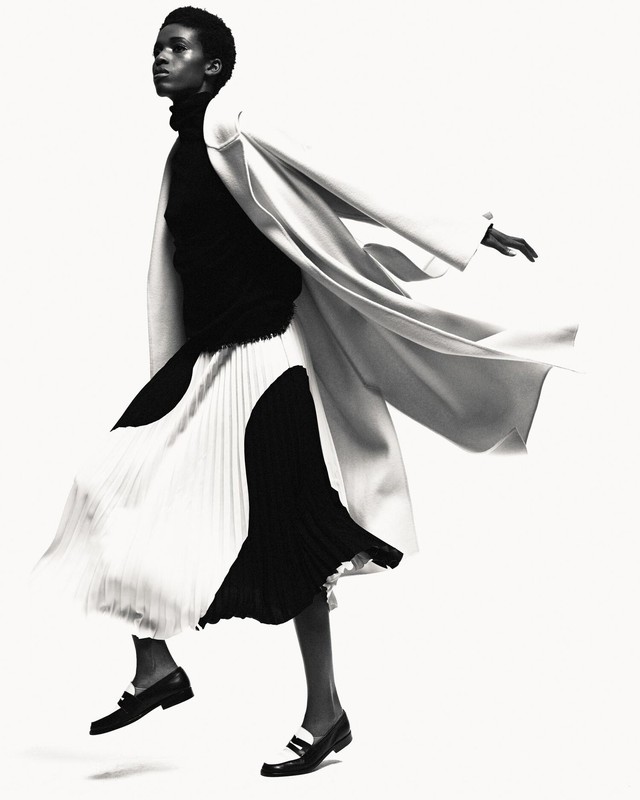 Monochrome fashion shot of a Black woman in a black turtleneck, flowing black and white skirt, coat, and loafers against a white backdrop.