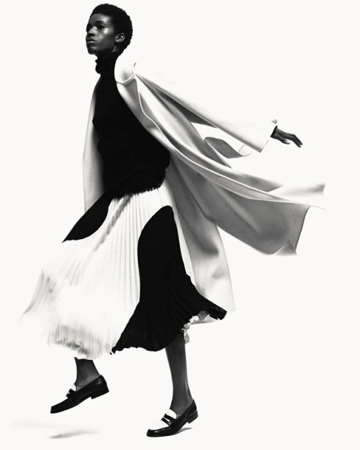 Monochrome fashion shot of a Black woman in a black turtleneck, flowing black and white skirt, coat, and loafers against a white backdrop.
