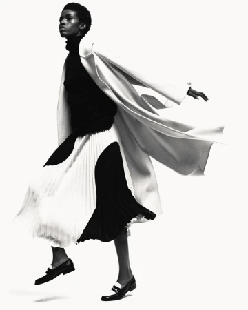 Monochrome fashion shot of a Black woman in a black turtleneck, flowing black and white skirt, coat, and loafers against a white backdrop.