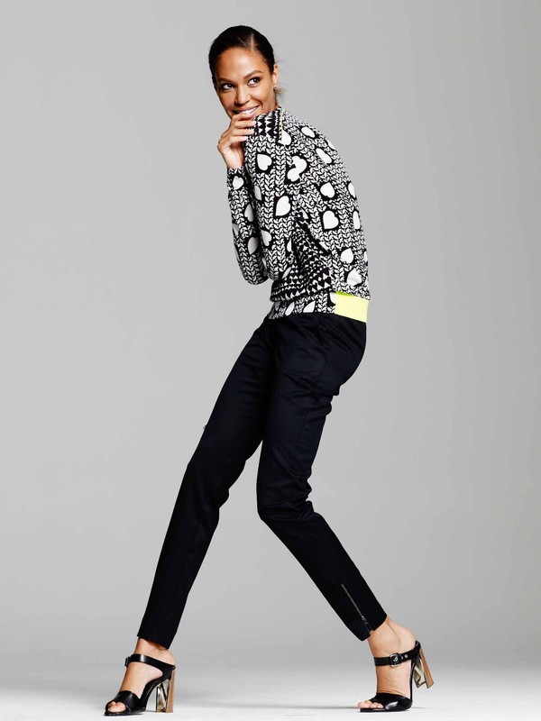 A full-length shot of a woman in stylish patterned sweater, dark trousers, and heeled sandals, posing in a light studio setting.