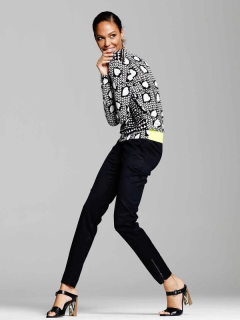 A full-length shot of a woman in stylish patterned sweater, dark trousers, and heeled sandals, posing in a light studio setting.