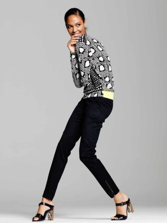 A full-length shot of a woman in stylish patterned sweater, dark trousers, and heeled sandals, posing in a light studio setting.