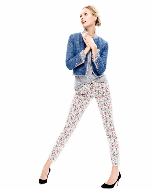 A full-length studio shot of a fashion model posing in a blue denim jacket, floral pants, and black heels against a bright white backdrop.