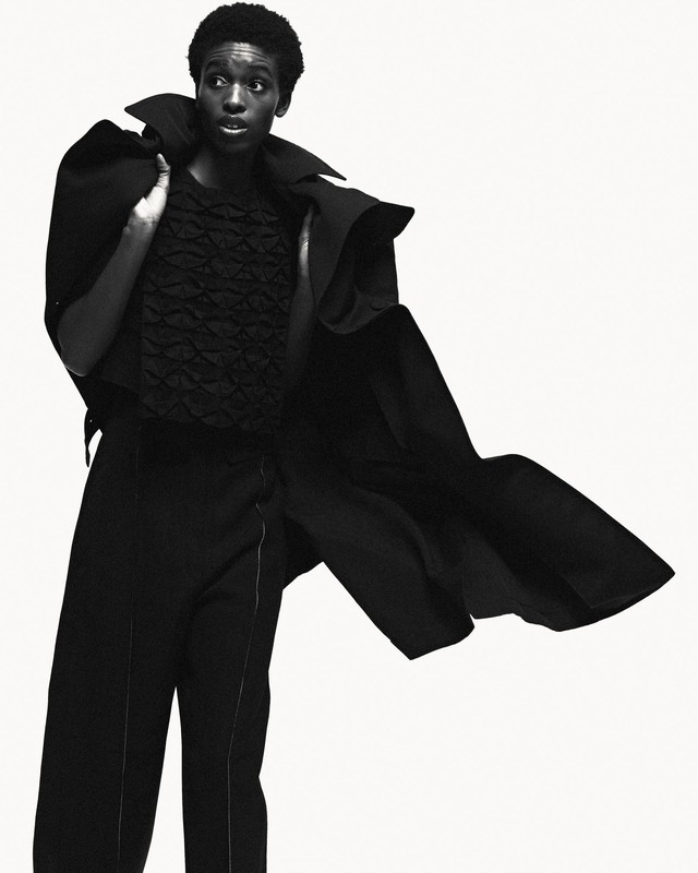 Fashion portrait of a Black woman wearing dramatic black coat and pants against white backdrop in a photo studio, editorial-style image.