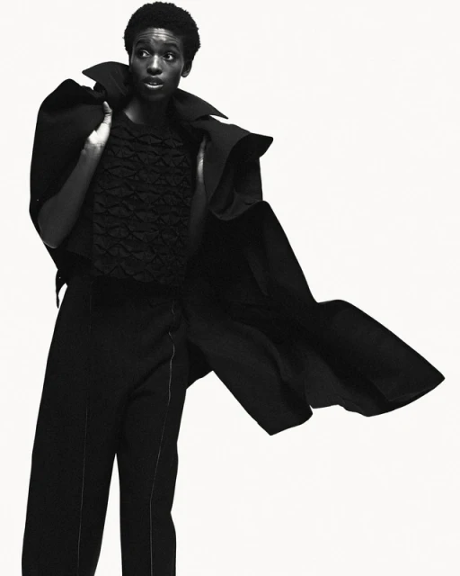 Fashion portrait of a Black woman wearing dramatic black coat and pants against white backdrop in a photo studio, editorial-style image.