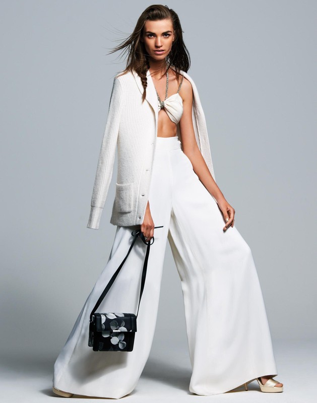 Full-length studio shot of a fashion model wearing all white outfit with a floral purse against a light gray backdrop. 
