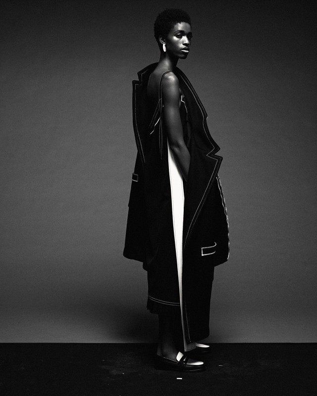 Elegant Black woman poses in a black and white fashion shot, wearing avant-garde coat and dress with white seam details on a gray background.