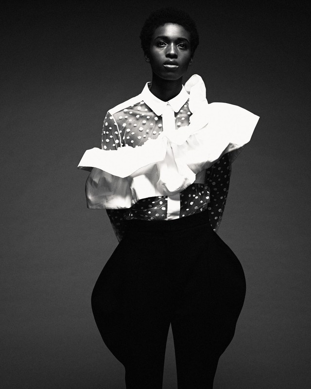Black and white portrait of an African American female model in a polka dot blouse with white ruffled collar, posing against a grey background.
