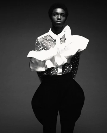 Black and white portrait of an African American female model in a polka dot blouse with white ruffled collar, posing against a grey background.